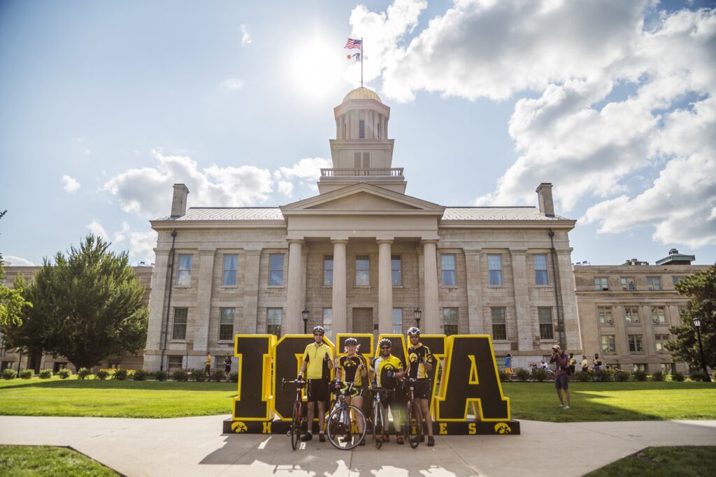 Four Iowa Hawkeye bicyclists standing in front of the Iowa Hawkeyes sign in front of the Old Capitol building in Downtown Iowa City.